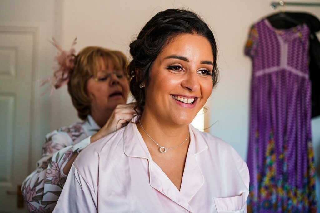 A woman smiles brightly as she gets ready, wearing a light pink pajama top and a necklace. Another woman, dressed in floral, stands behind her, assisting with her hair. In the background, a purple dress with a floral pattern hangs on the wall. Image by Documentary Wedding Photographer Lewis Bishop Photography.