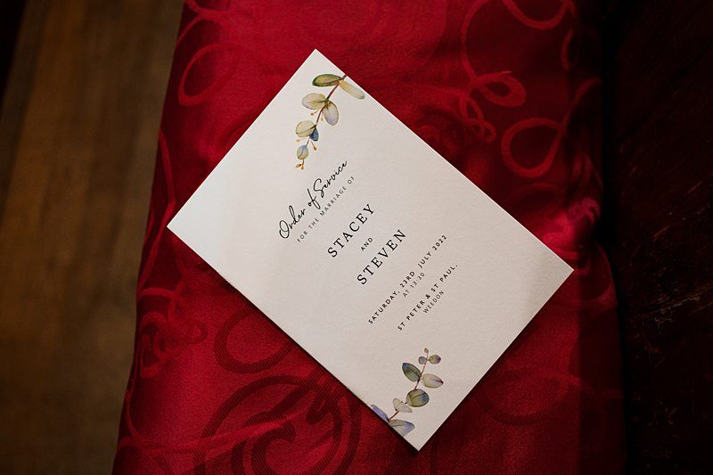 A white wedding program featuring floral decor and text reading "Order of Service for the Wedding of Stacey and Steven. Saturday, July 15, 2017. St. Peter's Church Hall" is placed on a red, patterned fabric background. Image by Documentary Wedding Photographer Lewis Bishop Photography.