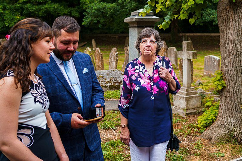 A man and two women stand together in a cemetery. The man, wearing a blue suit, is holding a phone. One woman, in a floral dress, looks at him while the other, in a floral blouse, looks at the camera with a solemn expression. Tombstones are visible in the background. Image by Documentary Wedding Photographer Lewis Bishop Photography.