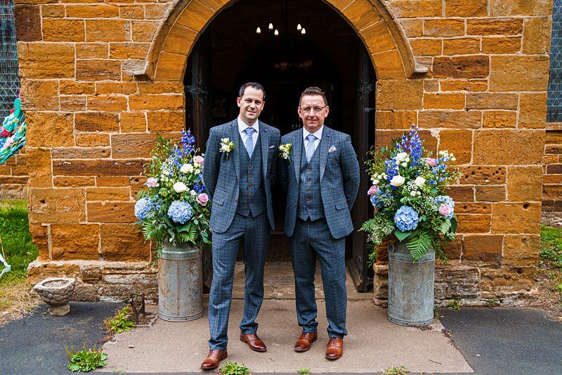 Two men in matching blue-checkered suits stand side by side in front of a rustic brick church entrance adorned with large floral arrangements. They appear well-dressed for a formal occasion, possibly a wedding. Image by Documentary Wedding Photographer Lewis Bishop Photography.