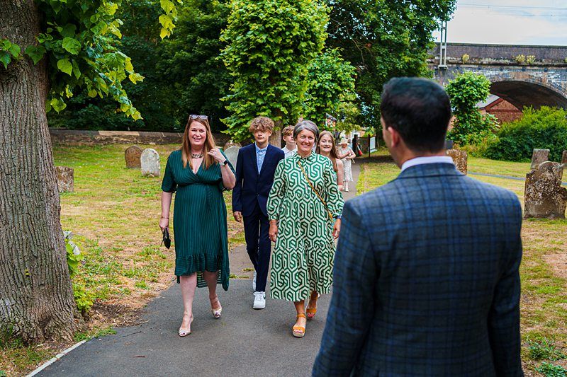 A group of people walking on a path through a park with green grass and trees. A man in a suit approaches the group while a woman in a green dress, a young man in a navy suit, and another woman in a patterned dress smile and walk towards him. Image by Documentary Wedding Photographer Lewis Bishop Photography.