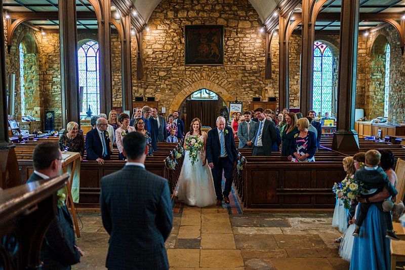 A bride, escorted by an older man, walks down the aisle of a rustic church filled with guests. Stained glass windows and wooden pews are visible. A man in a suit, likely the groom, stands at the altar awaiting her arrival. Bridesmaids and flower girls are on the right. Image by Documentary Wedding Photographer Lewis Bishop Photography.