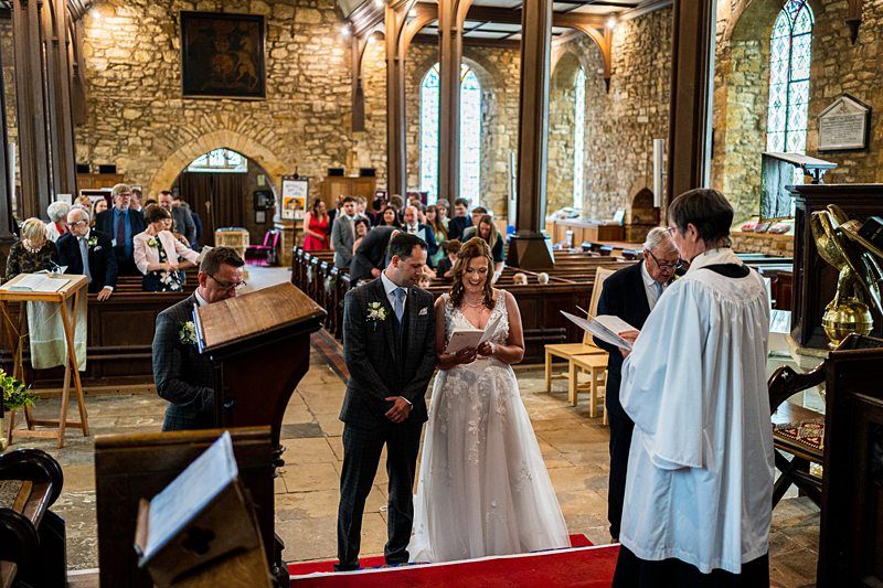 A bride and groom stand together at the altar in a stone church, accompanied by a priest who reads from a book. Guests are seated in wooden pews and some are standing, all dressed formally. Stained glass windows and wooden arches adorn the church interior. Image by Documentary Wedding Photographer Lewis Bishop Photography.