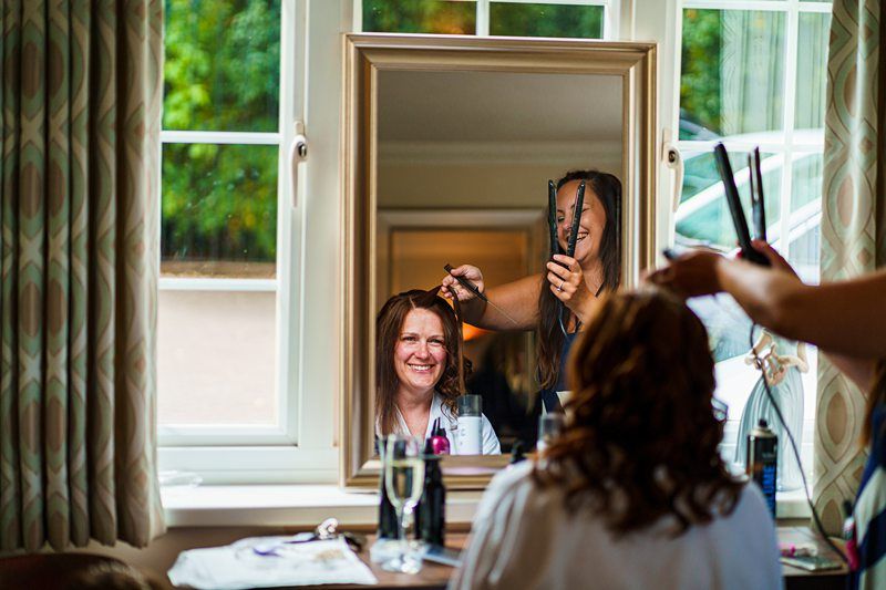 A woman smiles while sitting in front of a mirror as a hairstylist uses a curling iron to style her hair. The scene takes place in a well-lit room with a large window in the background. A glass of champagne and various hair products are visible on the table. Image by Documentary Wedding Photographer Lewis Bishop Photography.