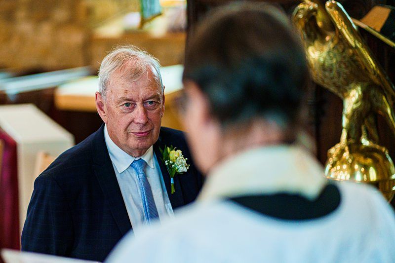 A man with gray hair and wearing a dark suit, light blue tie, and white boutonniere, stands in a church, looking towards a person in a white robe (the back of which is visible). There is a golden eagle lectern in the background.
. Image by Documentary Wedding Photographer Lewis Bishop Photography.