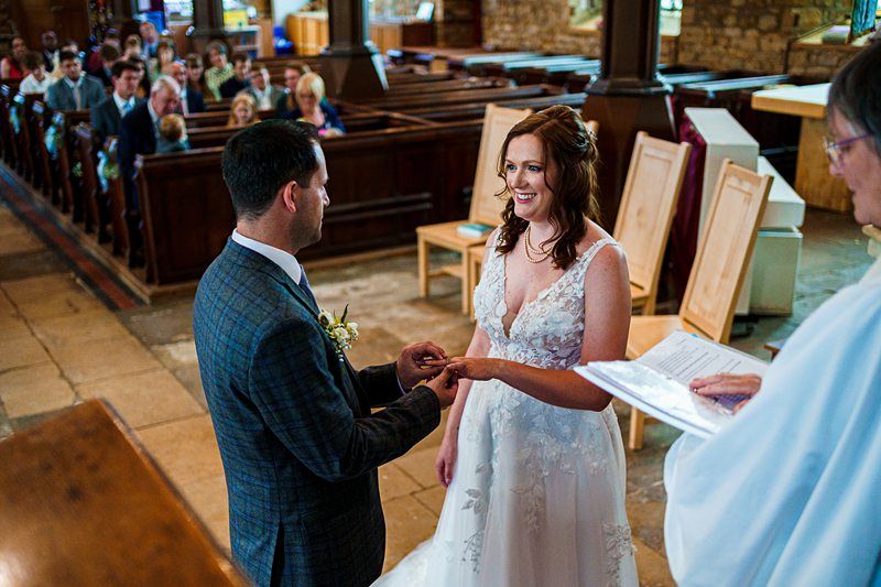 A couple stands at the altar exchanging rings during their wedding ceremony in a church. The bride is wearing a white lace dress, and the groom is in a blue checkered suit. Guests are seated in wooden pews. An officiant holding a book is partially visible on the right. Image by Documentary Wedding Photographer Lewis Bishop Photography.