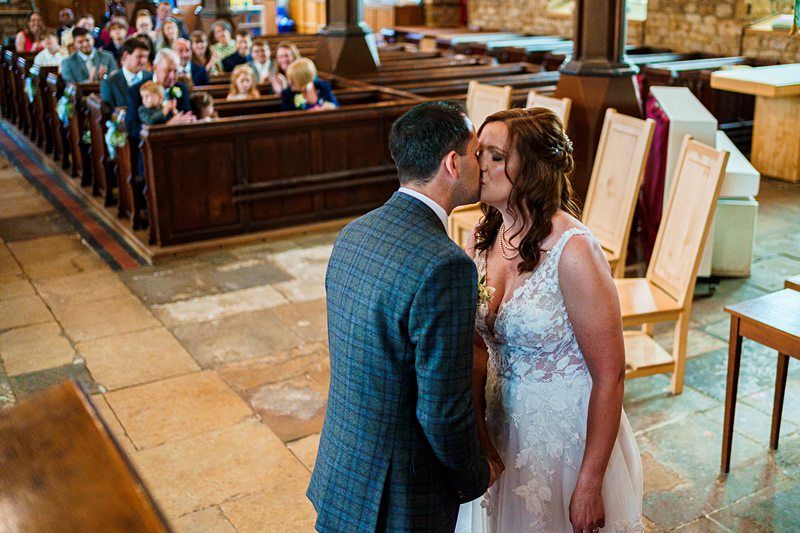 A bride and groom share a kiss inside a church during their wedding ceremony while guests sit in pews and watch. The bride wears a white wedding dress, and the groom is in a blue checkered suit. Image by Documentary Wedding Photographer Lewis Bishop Photography.