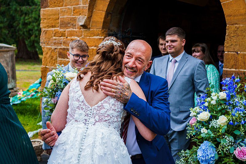 A bride in a white lace dress with floral headpiece hugs an older man in a blue suit and tie in front of a brick archway. Smiling guests, including a young boy with glasses and a woman in a green dress, surround them with colorful floral arrangements nearby. Image by Documentary Wedding Photographer Lewis Bishop Photography.