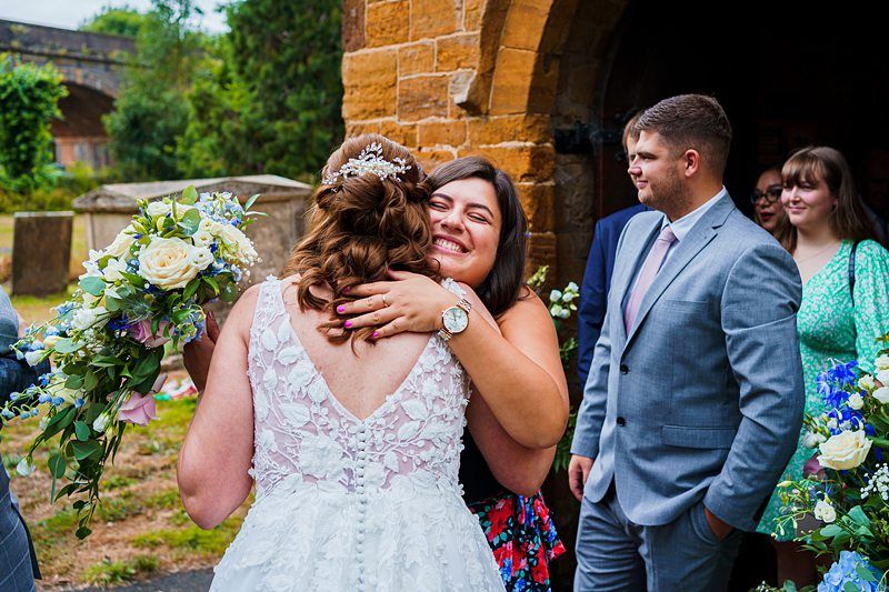 A bride in a lace wedding dress is hugging a smiling woman while holding a bouquet. They are outdoors, in front of a stone building with guests around them, including a man in a gray suit and a woman in a green dress. The atmosphere is joyful and celebratory. Image by Documentary Wedding Photographer Lewis Bishop Photography.