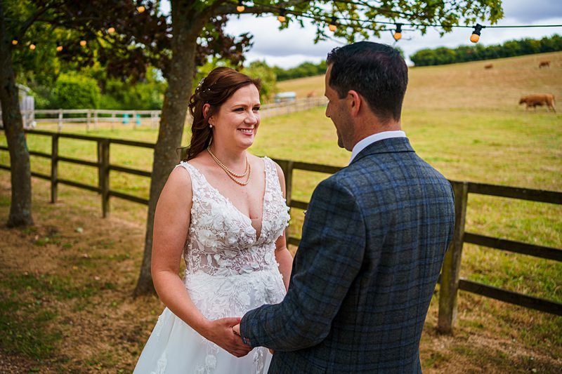 A bride and groom stand outdoors in a rural setting, holding hands and smiling at each other. The bride is wearing a white dress with floral details, and the groom is in a checkered suit. Behind them are trees, a wooden fence, and fields with grazing cows. Image by Documentary Wedding Photographer Lewis Bishop Photography.