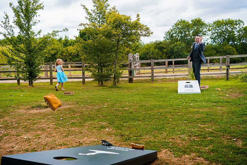 A man in a suit and a young girl in a blue dress play cornhole outside on a grassy field with trees and a wooden fence in the background. The man is tossing a bean bag, and one bean bag is mid-air. The game boards are positioned opposite each other. Image by Documentary Wedding Photographer Lewis Bishop Photography.