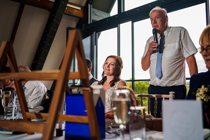 A man in a white shirt and blue tie is speaking into a microphone at a wedding reception. A bride in a white dress is seated next to him, smiling. Other guests are partially visible at the table, which is set with glasses and decorations. Image by Documentary Wedding Photographer Lewis Bishop Photography.