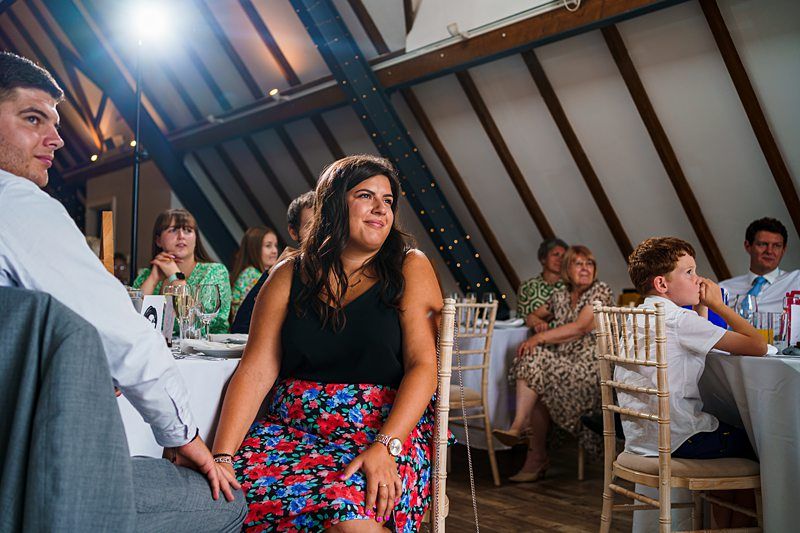 A group of people sitting at round tables in a room with a high ceiling and exposed wooden beams. Some are looking towards the front with expressions of interest and engagement. The atmosphere appears to be relaxed and casual, possibly during an event or gathering. Image by Documentary Wedding Photographer Lewis Bishop Photography.