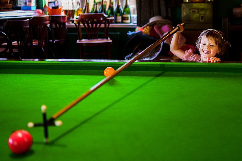 A young child, smiling brightly, uses a long cue to play on a green snooker table. The child stands on the side of the table, aiming at a red ball. Background elements include chairs and various items, creating a cozy indoor setting. Image by Documentary Wedding Photographer Lewis Bishop Photography.
