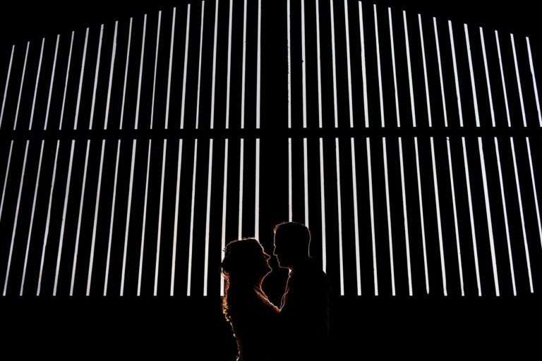 Silhouetted couple standing close to each other, with vertical white light beams creating a dramatic background against the dark setting. The vertical lines form an abstract, almost geometric backdrop, highlighting the couple's intimate moment. Image by Documentary Wedding Photographer Lewis Bishop Photography.