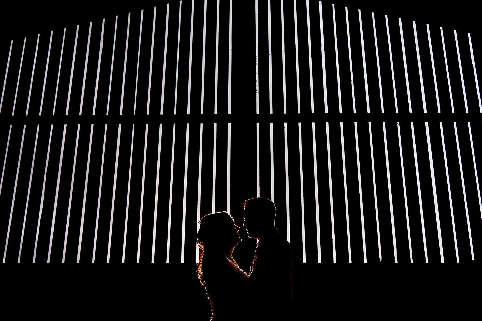 Silhouetted couple standing close to each other, with vertical white light beams creating a dramatic background against the dark setting. The vertical lines form an abstract, almost geometric backdrop, highlighting the couple's intimate moment. Image by Documentary Wedding Photographer Lewis Bishop Photography.