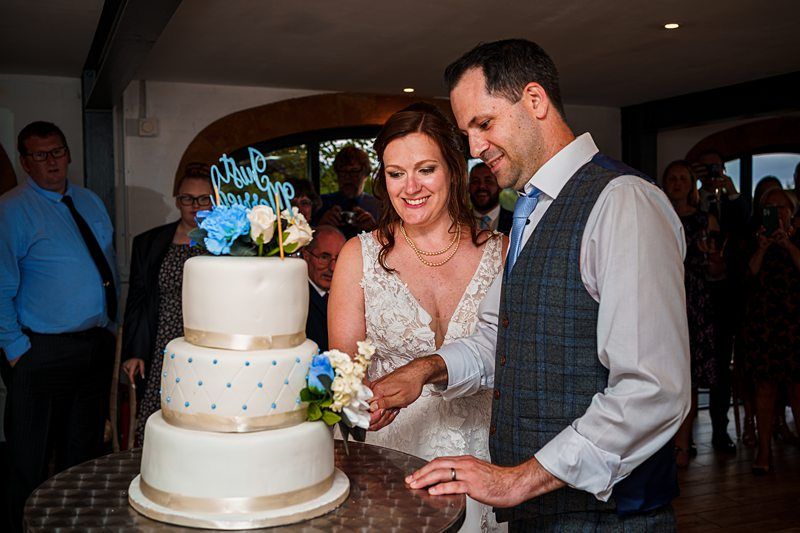 A bride in a white lace wedding dress and a groom in a white shirt with a vest cut a tiered wedding cake adorned with blue and white flowers. Guests in the background watch and take pictures in a warmly lit reception area. Image by Documentary Wedding Photographer Lewis Bishop Photography.