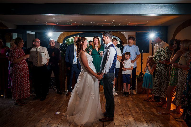A bride and groom hold hands and gaze at each other on a wooden floor, surrounded by smiling guests. The scene is lit by spotlights, and everyone appears to be enjoying a celebration, likely a wedding reception. Image by Documentary Wedding Photographer Lewis Bishop Photography.