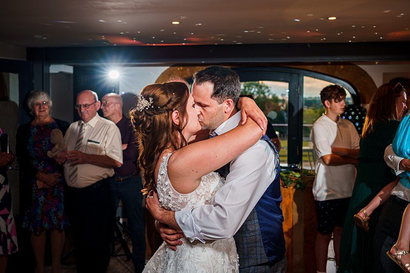 A bride in a white lace wedding dress and a groom in a blue vest embrace each other while dancing, surrounded by guests watching them. The room is warmly lit with a visible window and some decorative lights in the background. Image by Documentary Wedding Photographer Lewis Bishop Photography.