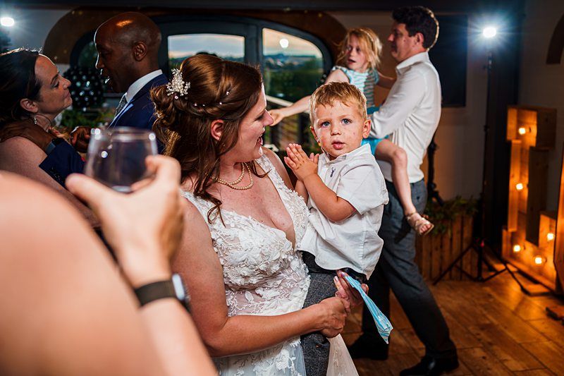 A bride in a white wedding dress holds a young boy on a dance floor. Guests in the background, including another child being held by an adult, are dancing and socializing. The setting appears to be a lively wedding reception with wooden floors and large windows. Image by Documentary Wedding Photographer Lewis Bishop Photography.
