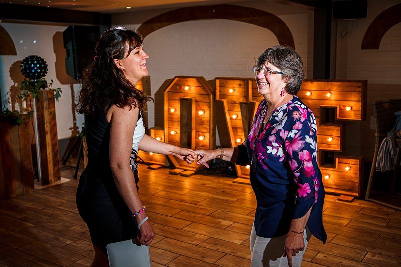 Two women smile and hold hands while dancing in a warmly lit room with a large illuminated "LOVE" sign in the background. One woman is wearing a black and white dress, and the other is in a floral blouse with white pants. The atmosphere is joyful. Image by Documentary Wedding Photographer Lewis Bishop Photography.