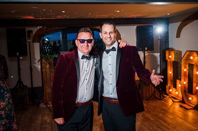 Two men in matching red velvet blazers, black bow ties, and white shirts stand arm in arm, smiling at the camera. One wears sunglasses, and they are in a room lit by a spotlight and large decorative letters. Wood flooring and rustic decor are visible in the background. Image by Documentary Wedding Photographer Lewis Bishop Photography.