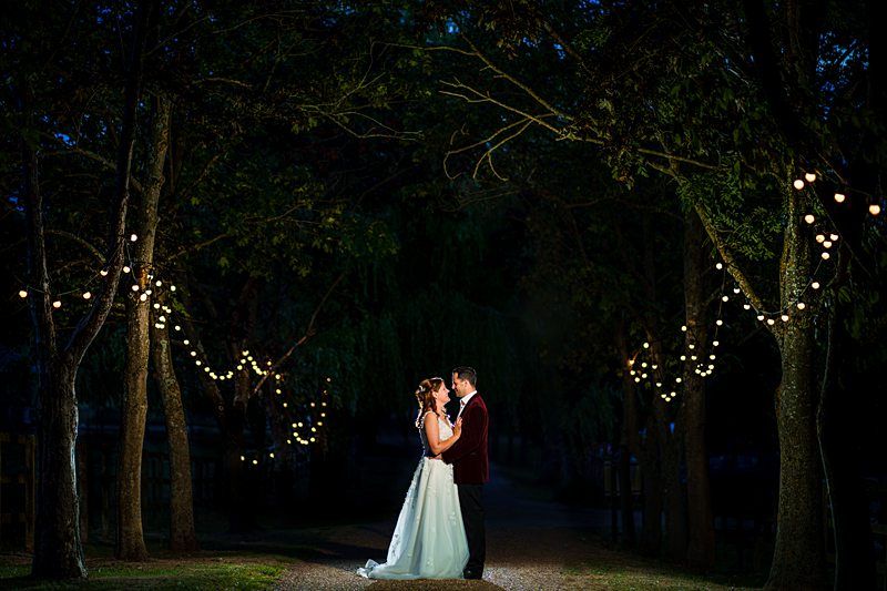 A bride in a white dress and a groom in a dark suit are standing close together, gazing into each other's eyes. They are on a tree-lined path adorned with string lights. The background is dark, emphasizing the couple and the illuminated path. Image by Documentary Wedding Photographer Lewis Bishop Photography.