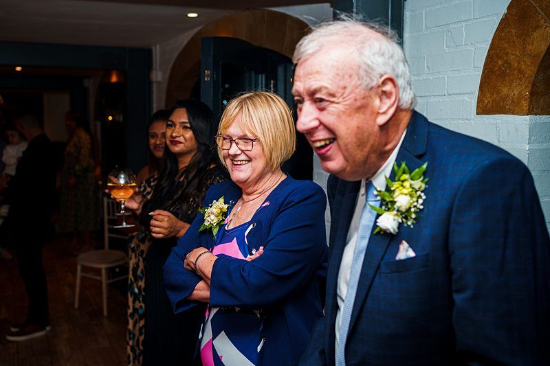 A smiling older couple, dressed formally with floral boutonnieres, stand beside each other at a social gathering. The woman has blonde hair and glasses, while the man has gray hair. Behind them, several other people are also smiling and conversing. Image by Documentary Wedding Photographer Lewis Bishop Photography.