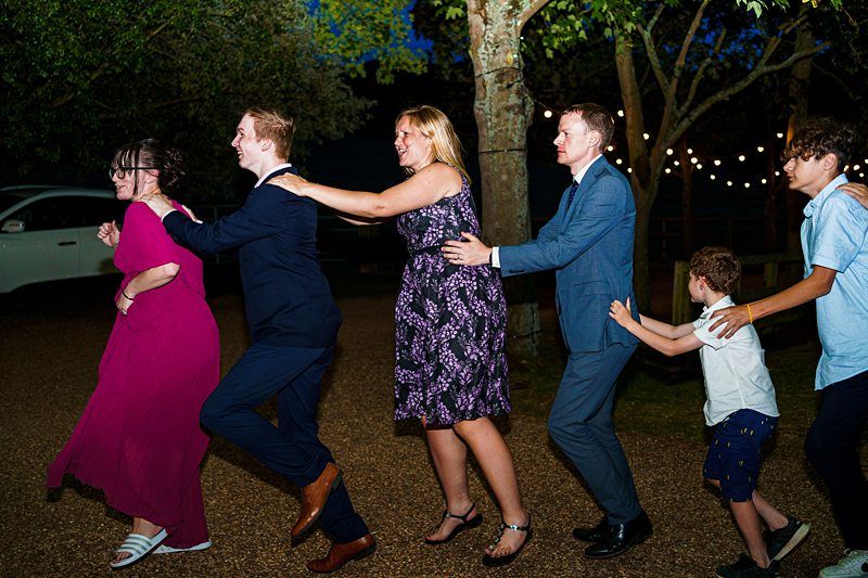 A group of people, dressed in formal and semi-formal attire, are dancing in a conga line outdoors at night. The scene is illuminated by string lights, with a backdrop of trees. Everyone appears to be smiling and having a good time. Image by Documentary Wedding Photographer Lewis Bishop Photography.