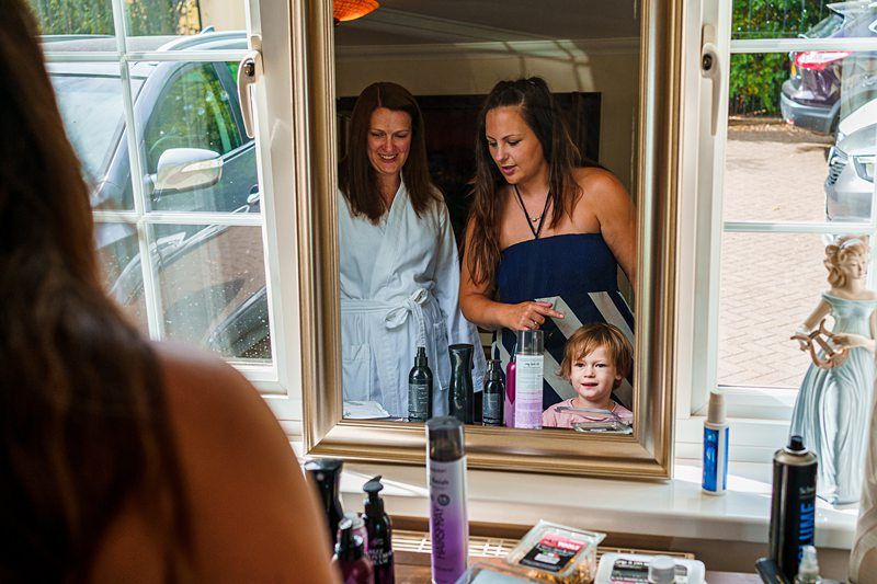 A woman in a white robe and another in a navy dress smile while looking at a young child through a mirror. The vanity table is cluttered with various beauty products, and there is a parked car seen through the window. Image by Documentary Wedding Photographer Lewis Bishop Photography.