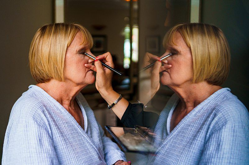 An older woman with short blonde hair is having her makeup done by a person with a brush. She is wearing a light blue robe and her reflection is visible in the mirror in front of her. The background is slightly blurred, featuring a soft-lit room. Image by Documentary Wedding Photographer Lewis Bishop Photography.