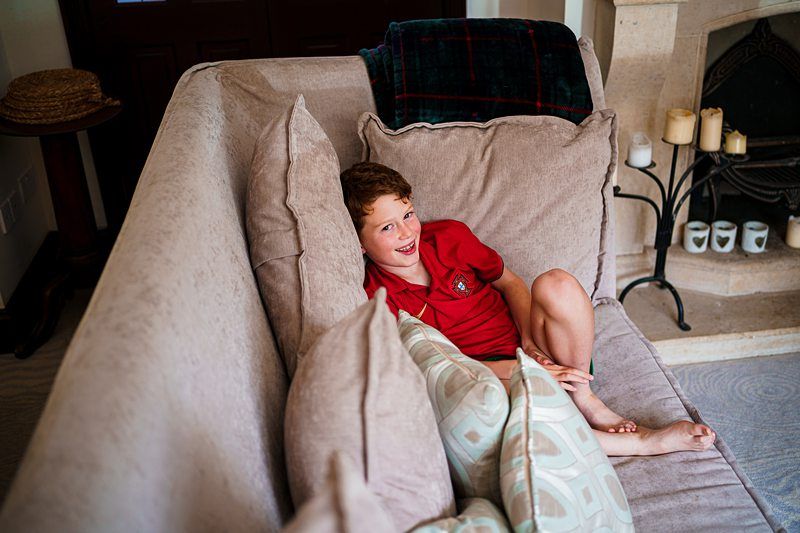 A young boy with red hair wearing a red polo shirt is lounging on a beige sofa among large, soft cushions. He is smiling and looking at the camera. In the background, there is a fireplace with candles and a mantel with a folded blanket on it. Image by Documentary Wedding Photographer Lewis Bishop Photography.