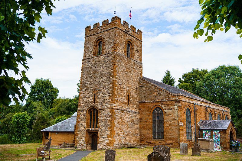 An old stone church with a tall rectangular tower and clock, surrounded by trees and gravestones. The church building is made of weathered stone and features arched windows. The sky is partly cloudy, and the scene is framed by green foliage. Image by Documentary Wedding Photographer Lewis Bishop Photography.