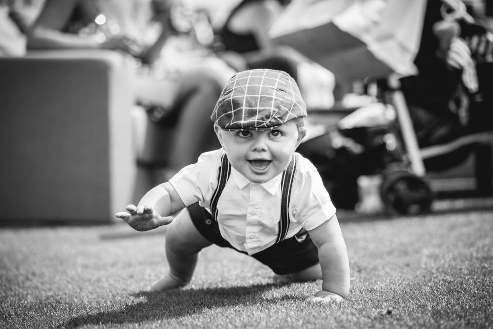 A black-and-white photo of a baby wearing a checked flat cap, a white shirt, and dark shorts with suspenders, crawling on grass. The baby has a cheerful expression with an open mouth. In the blurred background, there are people and a stroller. Image by Documentary Wedding Photographer Lewis Bishop Photography.