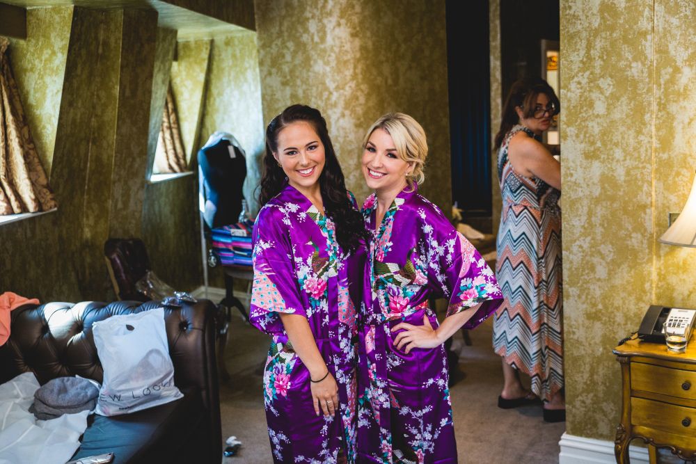Two women in floral purple robes stand smiling in a warmly lit room with textured golden walls. A dresser with a lamp and bags on a chair are on the left, and another woman stands in the background wearing a colorful dress, looking towards the camera. Image by Documentary Wedding Photographer Lewis Bishop Photography.