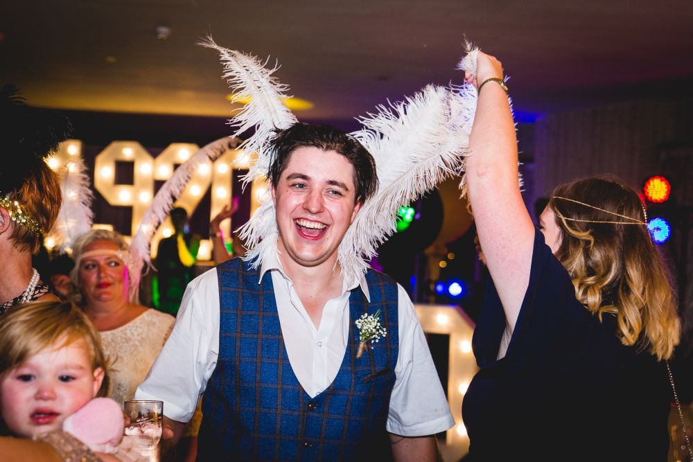 A man in a white shirt and blue vest is laughing at a festive event. He is wearing a headpiece with large white feathers, and a woman next to him is holding her arm up, also with white feathers. People in the background are smiling, and a child is in the foreground. Image by Documentary Wedding Photographer Lewis Bishop Photography.