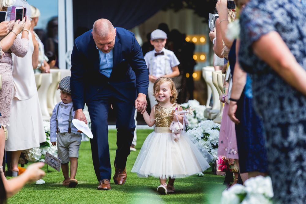 A smiling young girl in a white and gold dress holds hands with an older man in a suit as they walk down a grassy aisle, surrounded by people clapping and taking photos. Another young boy in suspenders and a flat cap follows behind, holding a hat. Image by Documentary Wedding Photographer Lewis Bishop Photography.