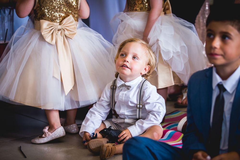 A young boy dressed in white with suspenders and a bow tie sits on the floor, looking upward. Other children, dressed in fancy attire, stand nearby. One girl wears a dress with a tulle skirt and a large bow, while a boy in a blue suit sits on the right. Image by Documentary Wedding Photographer Lewis Bishop Photography.