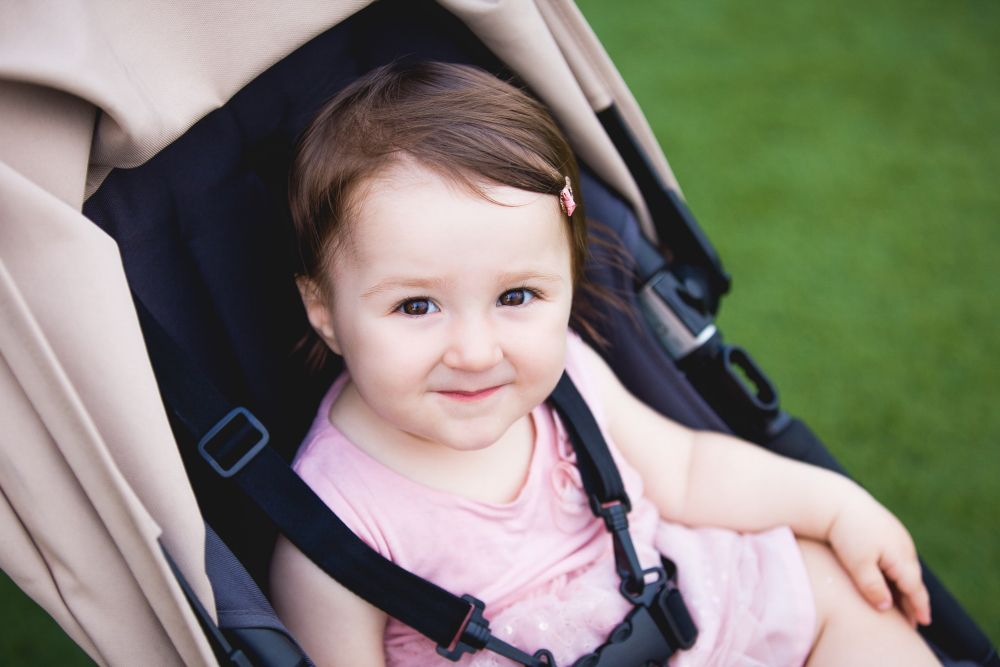 A smiling toddler with brown hair, adorned with a small pink hair clip, sits in a beige stroller on a green lawn. She is wearing a light pink sleeveless top and looking up at the camera with a happy expression. Image by Documentary Wedding Photographer Lewis Bishop Photography.