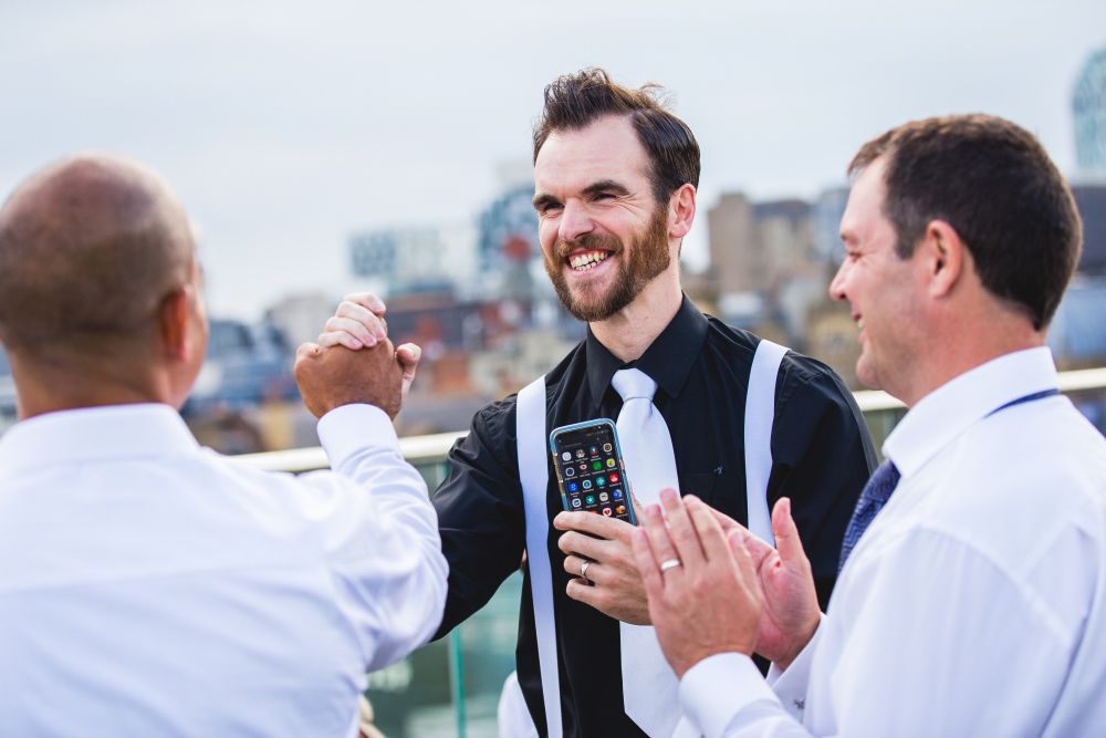 Three men in formal attire share a joyful moment outdoors. The man in the center, holding a smartphone, smiles and clasps hands with one of the men while the other man claps. The background shows a cityscape with buildings under a cloudy sky. Image by Documentary Wedding Photographer Lewis Bishop Photography.