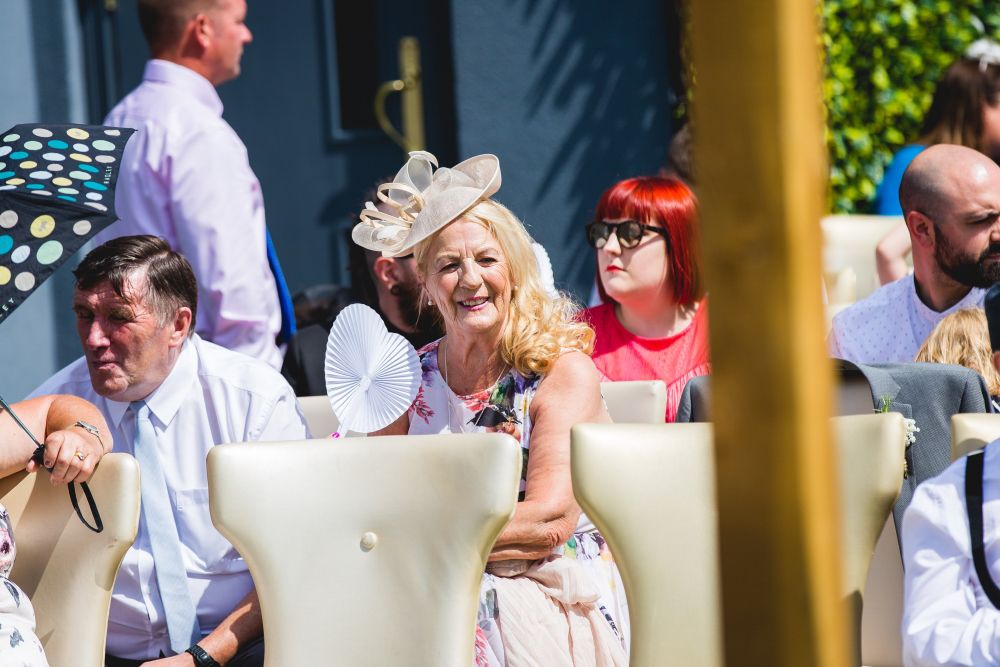 A crowd of people dressed in semi-formal attire sits outdoors during a sunny day. A smiling woman with wavy blonde hair, wearing a floral dress and a large hat, holds a hand fan. Other attendees, including a man beside her and a woman with red hair, are seated nearby. Image by Documentary Wedding Photographer Lewis Bishop Photography.