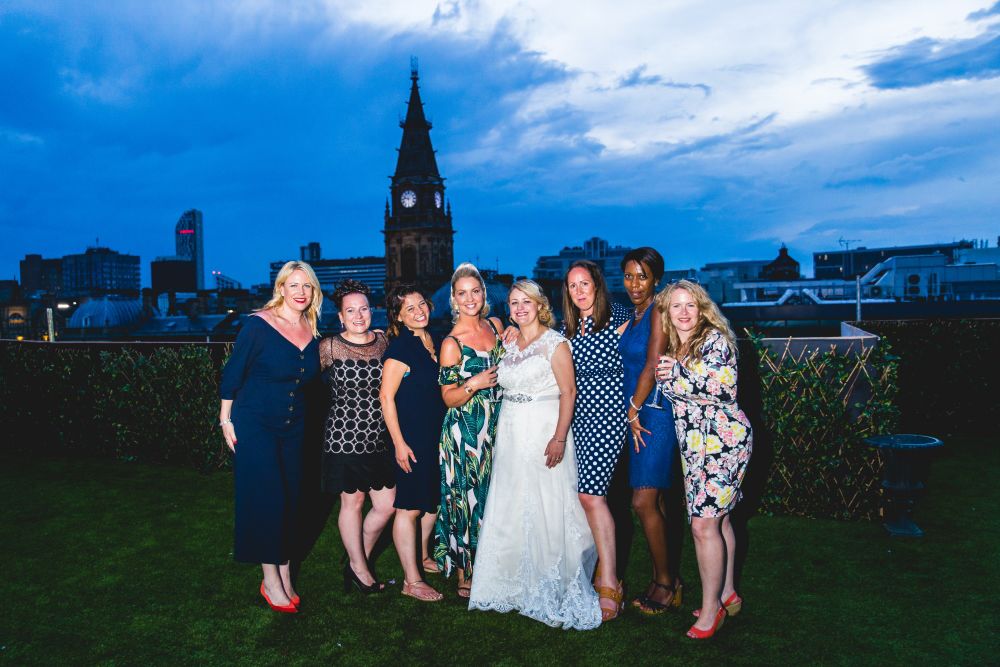 A group of eight women, including one in a wedding gown, pose for a photo on a grassy rooftop with a city skyline and clock tower in the background. The sky is blue with scattered clouds, and the women are wearing a variety of dresses, smiling at the camera. Image by Documentary Wedding Photographer Lewis Bishop Photography.