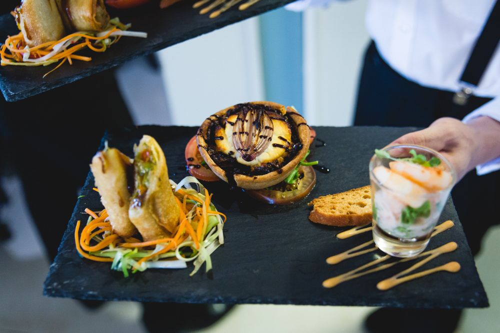 A person in formal attire carries a black slate plate with an assortment of gourmet appetizers. The plate includes stuffed pastry cups topped with balsamic drizzle, spring rolls on a bed of shredded vegetables, toast slices, and a glass with shrimp cocktail. Image by Documentary Wedding Photographer Lewis Bishop Photography.