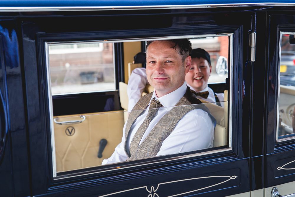 A man in a suit and tie sits in the front passenger seat of a classic blue car, smiling. A young boy in formal attire is visible behind him, also smiling. The car's window is partially rolled down, and the interior is cream-colored. Image by Documentary Wedding Photographer Lewis Bishop Photography.