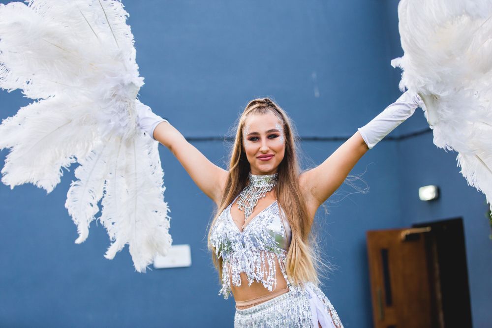 A woman wearing a silver sequined costume and white gloves holds large white feather fans above her head against a blue background. She has long blonde hair and is smiling at the camera. She appears to be dressed for a performance or celebration. Image by Documentary Wedding Photographer Lewis Bishop Photography.