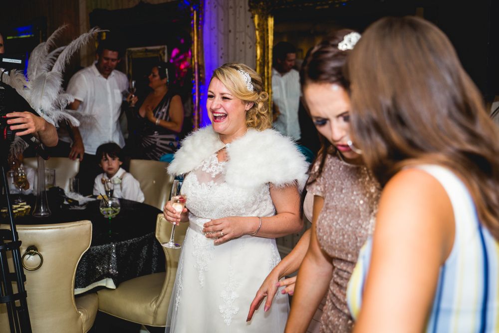 A bride in a white dress with a fur stole smiles and holds a champagne glass. She is surrounded by guests who are dancing and mingling. The background includes a little boy in a white shirt sitting at a table. The scene appears to be a festive celebration. Image by Documentary Wedding Photographer Lewis Bishop Photography.