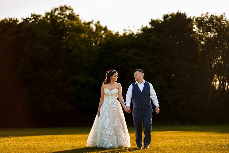 A couple dressed in wedding attire walks hand in hand through a grassy field. The bride wears a strapless, white gown with floral lace details, while the groom dons a dark vest, white shirt, and blue tie. Trees and sunset sky form the backdrop. Both appear happy and smiling. Image by Documentary Wedding Photographer Lewis Bishop Photography.