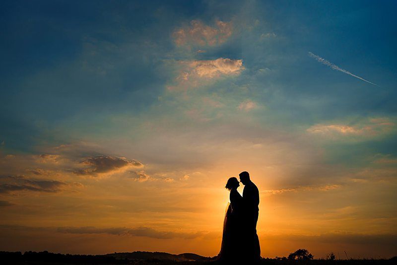 Silhouetted against a vibrant sunset, a couple embraces lovingly. The sky blends hues of orange, yellow, and blue while wispy clouds and a distant contrail add texture. The soft sunlight creates a serene and romantic atmosphere. Image by Documentary Wedding Photographer Lewis Bishop Photography.
