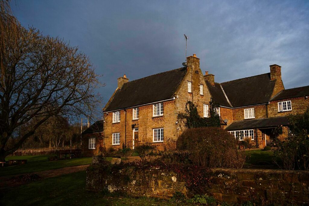 A traditional, two-story stone house with a gabled roof and multiple chimneys is set against a dark, cloudy sky. The property features a bare tree on the left, a low stone wall in front, and well-maintained gardens with shrubs and bushes. Image by Documentary Wedding Photographer Lewis Bishop Photography.