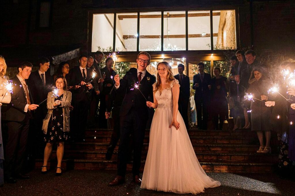 A bride and groom, both smiling, stand at the center of a staircase surrounded by wedding guests holding sparklers. The bride is wearing a white gown, and the groom is in a dark suit. The scene is lit warmly with sparklers and lights from a building behind them. Image by Documentary Wedding Photographer Lewis Bishop Photography.